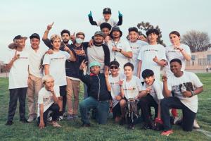 A baseball team coached by Markel and Martel Cooper poses together on a grassy field, wearing "Double Impakt" shirts and making number-one gestures