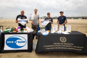 A GROUP OF PEOPLE STANDING BEHIND TABLES WITH ITEMS TO COLLECT TRASH AT THE BEACH