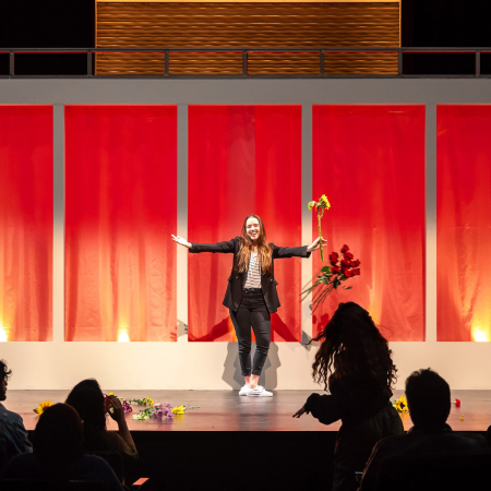 A performer stands onstage with arms outstretched, holding a yellow flower, while a silhouette in the audience throws flowers toward the stage.