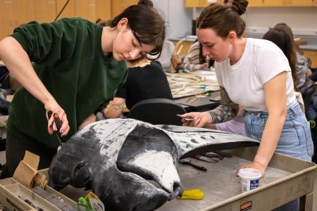 Two students work on a 3D-printed humpback whale mandible, using tools to smooth the surface in a marine biology classroom.