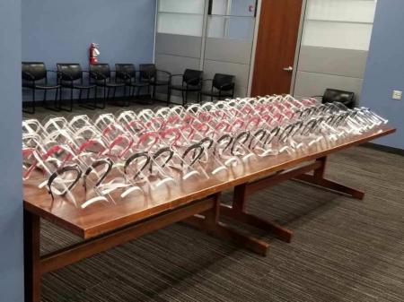 A large wooden table filled with rows of 3D-printed clear plastic face shields, arranged neatly in a conference room.