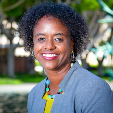 A person smiles at the camera, wearing a gray blazer and colorful necklace, standing outdoors.