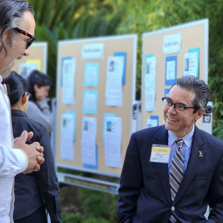 Two men speak in font of bulletin boards containing photographs and text.