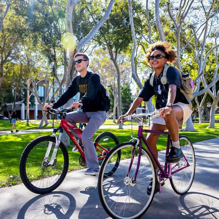 Two students ride bikes along a campus path surrounded by trees.