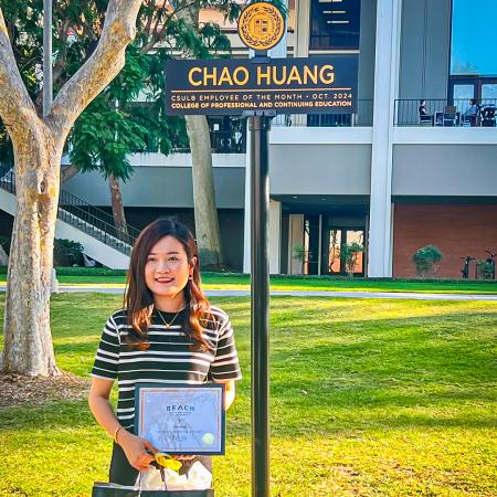 Chao Huang stands holding a certificate and bag under a sign bearing her name.