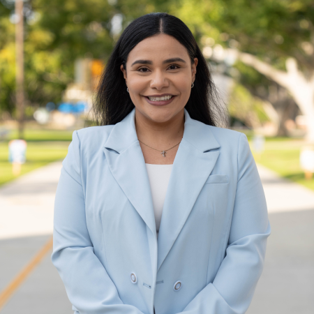 Celia Mejia smiles outdoors in a light blue blazer, with trees and a path in the background.