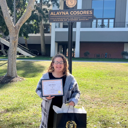 Alayna Cosores stands smiling outdoors holding a framed certificate and gift bag beneath a sign with her name on it. 