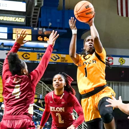 Beach basketball player Lovely Sonnier, wearing a yellow uniform, jumps for a shot while two Elon defenders in red attempt to block.