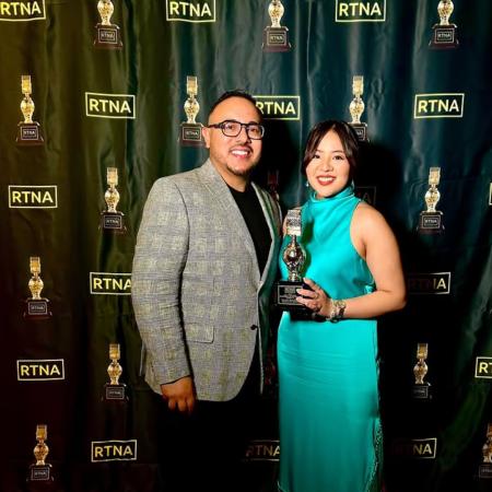 Student Raya Torres and broadcast journalism professor Jesús Ayala Two people stand in front of an RTNA step-and-repeat banner, with one person in dress holding a trophy shaped like a microphone