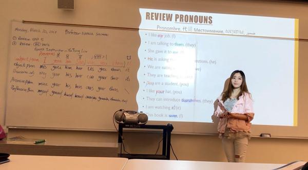 Young woman stands in front of a whiteboard, teaching pronouns in various languages