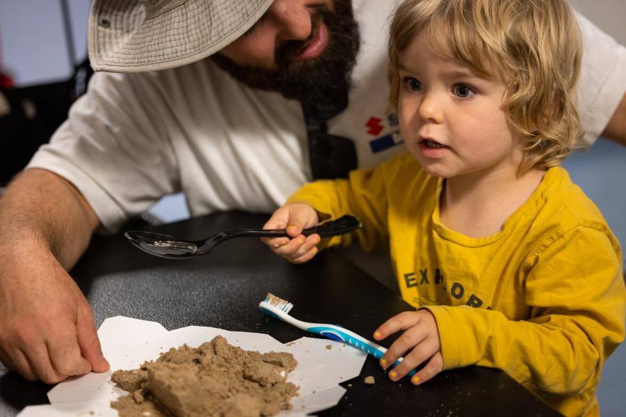 A toddler holds a toothbrush and looks for a fossil in a pile of sand while his dad speaks to him