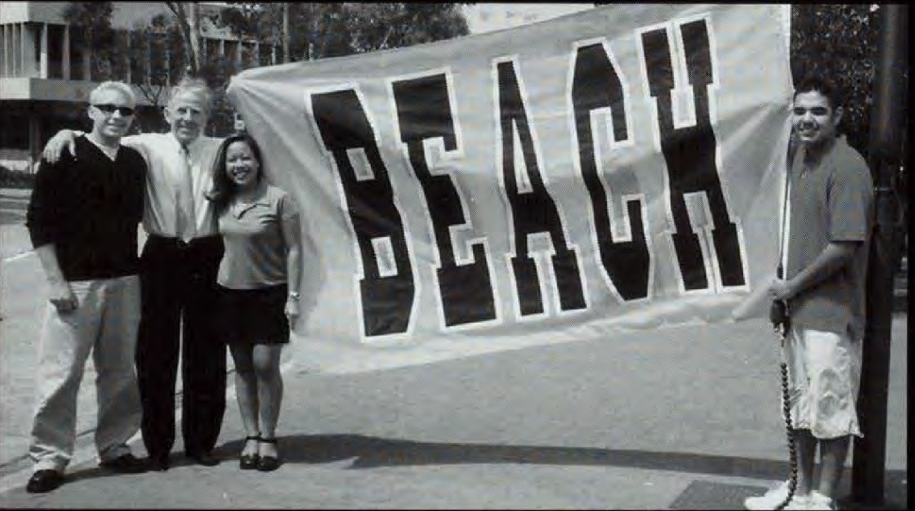 Four people stand by a large sign that reads "BEACH."