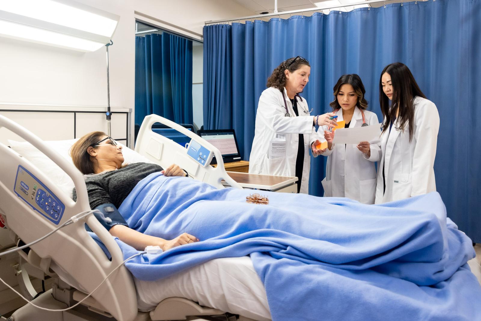 A woman acting as a patient lies in a hospital bed while two students in lab coats review documents with a faculty member in the School of Nursing.