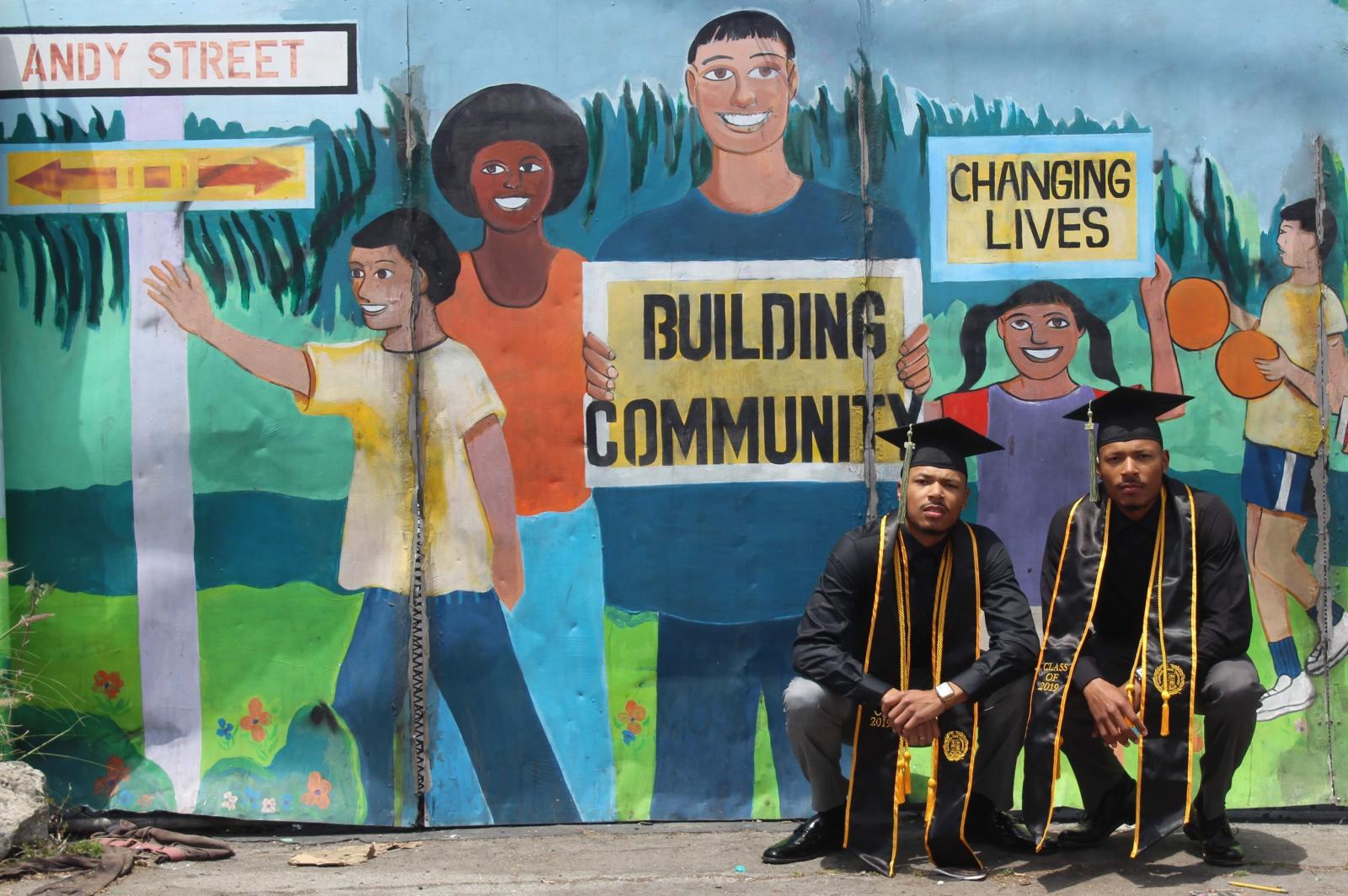 The Cooper twins, wearing CSULB caps and gowns, pose in front of a mural on on Andy Street where the grew up.